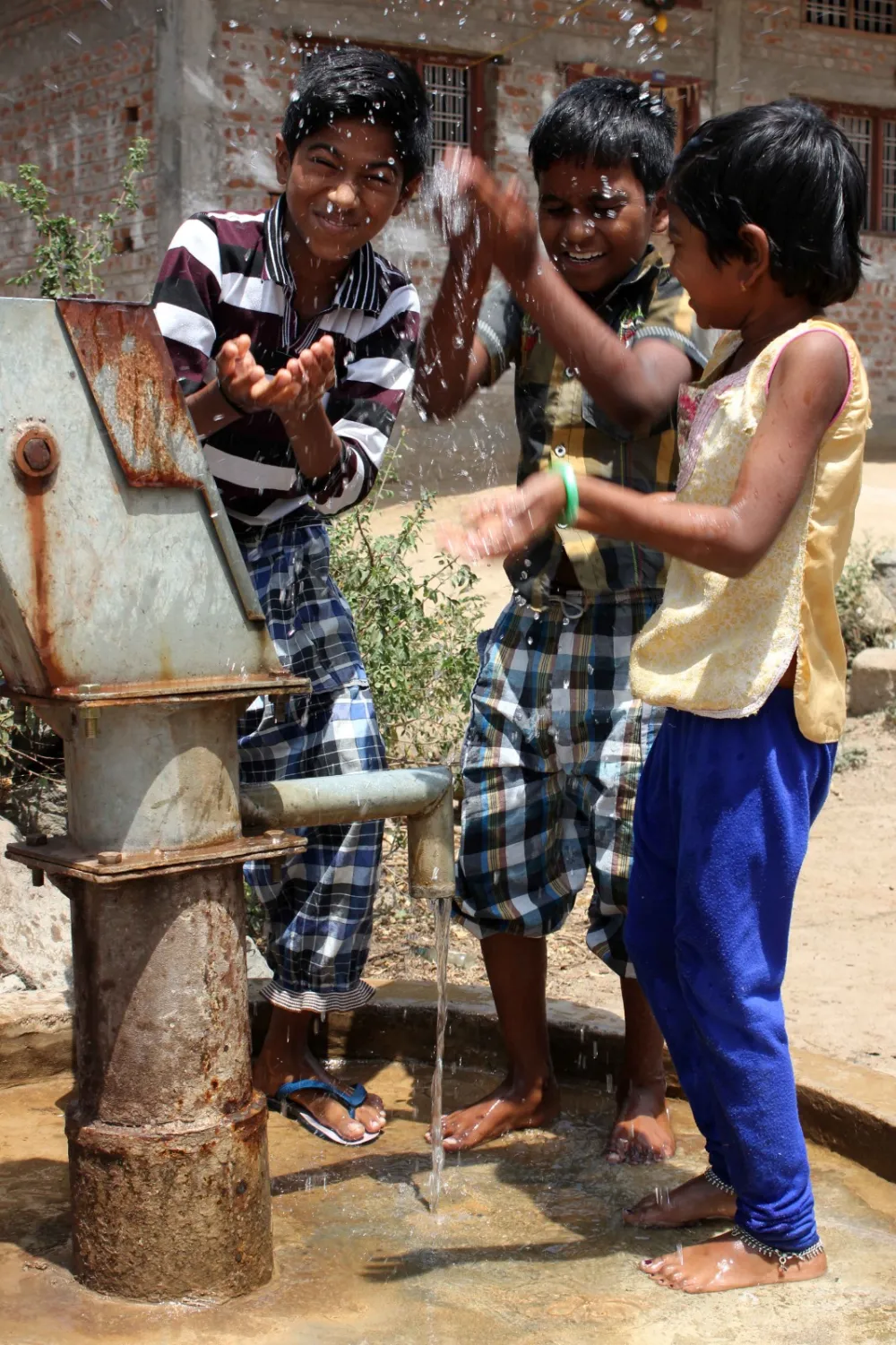 Three children laugh and splash in the water at a village hand pump.