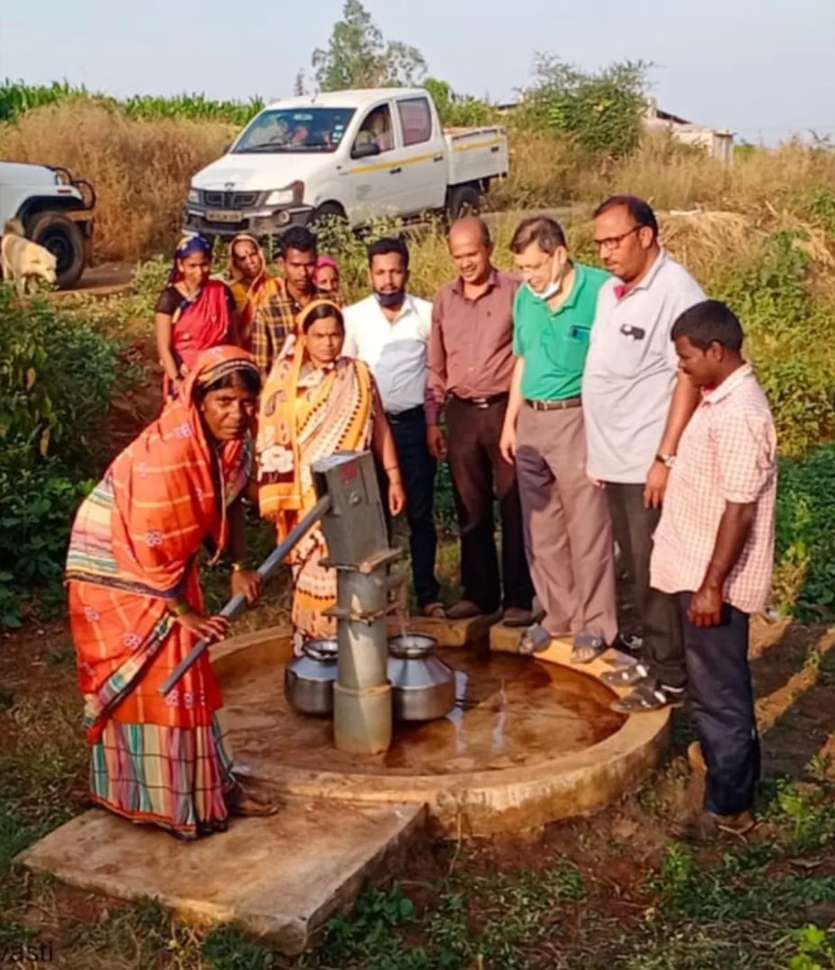 A woman draws water from a hand pump while villagers and officials look on at a rural water point in India.